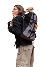 A woman wearing a gray and black dog backpack carrier with a small dog sitting inside, shown from the side against a plain white background.